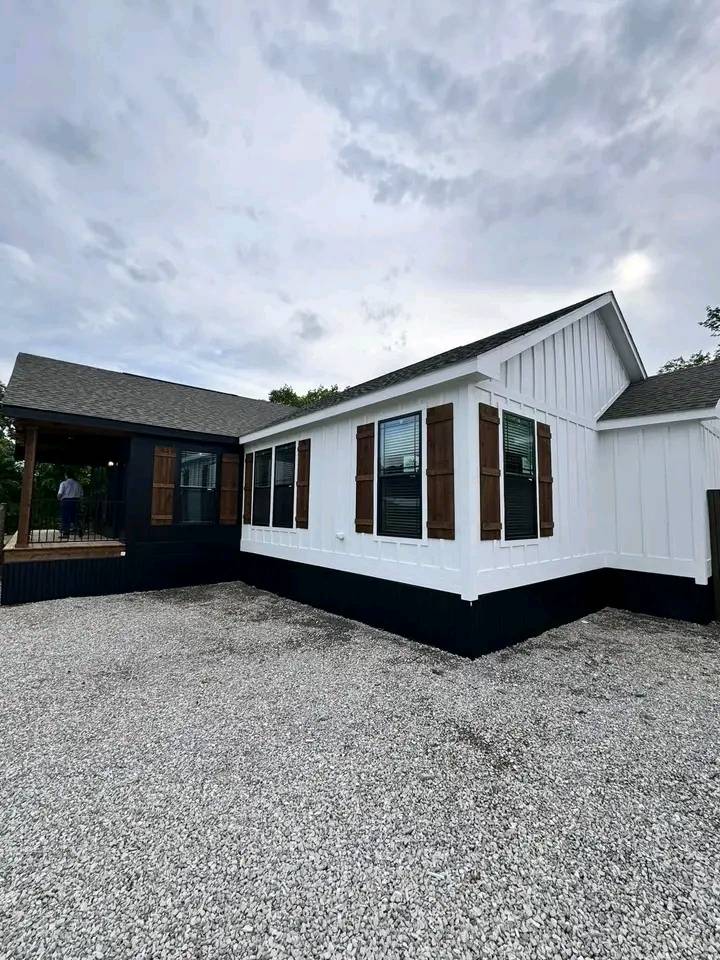 Modern white house with black trim, wooden shutters, and gravel driveway under an overcast sky. A person is visible on the wooden deck.