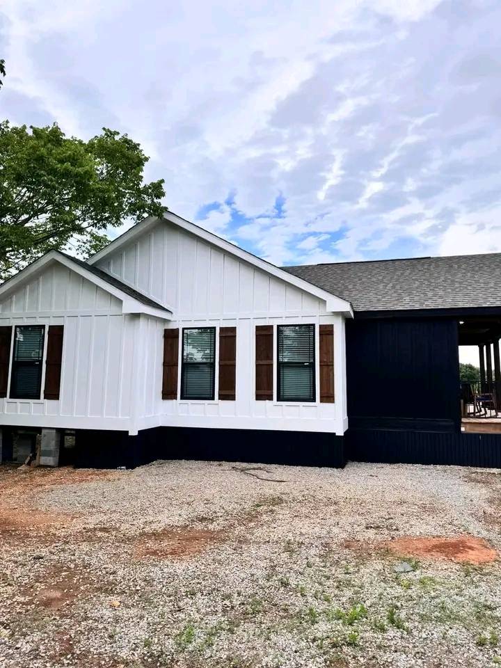 White and navy blue modern farmhouse with wooden shutters sits under a bright, partly cloudy sky. Gravel driveway and green tree add natural charm.