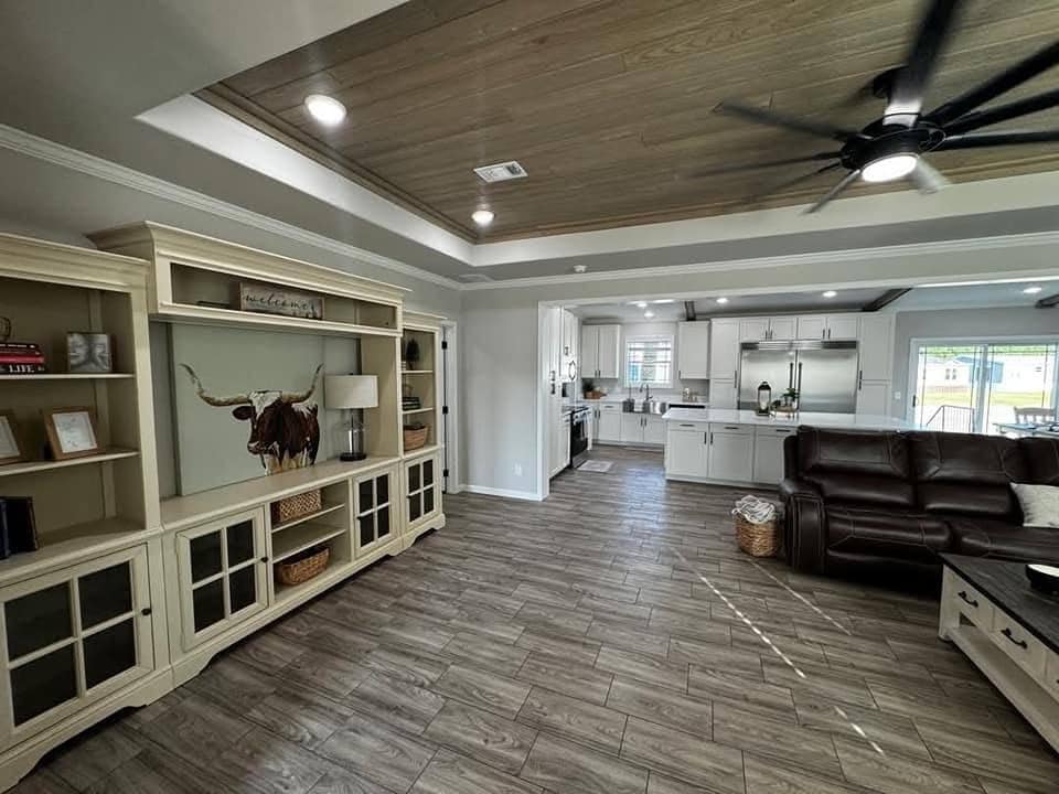 Modern living area with wooden floors, beige cabinets, and a leather couch. A ceiling fan spins above, and the kitchen is visible in the background.
