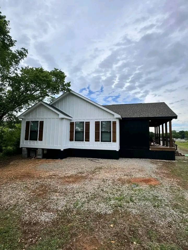 A modern farmhouse stands under a cloudy sky, featuring a white exterior with brown shutters and a black accent wall. A wraparound porch offers rustic charm.
