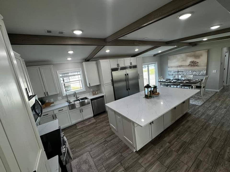 Spacious modern kitchen with white cabinets, large island, stainless steel appliances, and wooden beams on the ceiling. Dining area visible in the background.