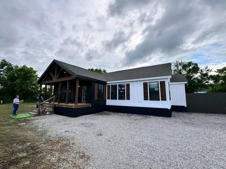Single-story house with black and white siding, a covered porch, and a gravel driveway. Two people stand outside under a cloudy sky. Calm atmosphere.