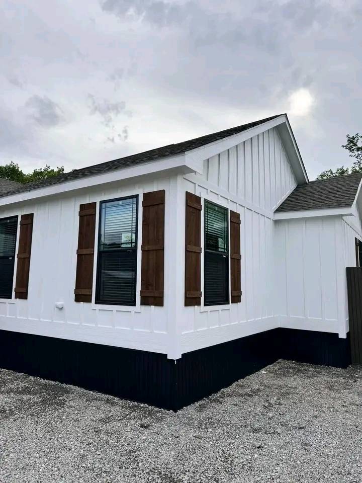 A modern, white house with vertical siding and dark brown shutters stands under a cloudy sky. Black trim accents the base, creating a clean contrast.