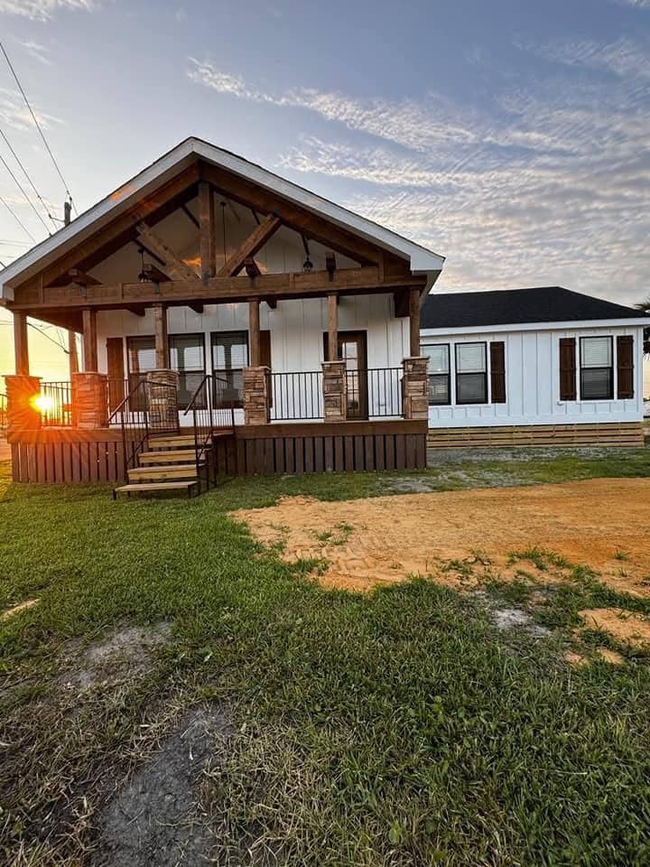 A cozy white farmhouse with a rustic wooden porch and stone pillars, bathed in warm sunset light. The sky is partly cloudy, creating a serene ambiance.
