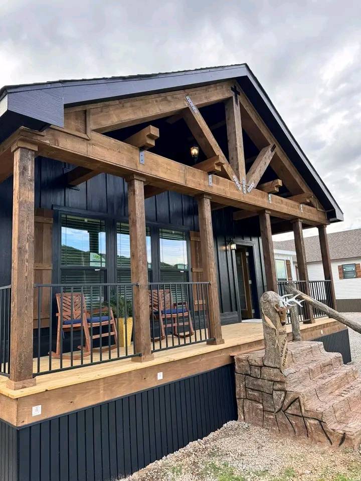 Cozy cabin with a wooden porch, two chairs, and a vintage water pump. Rustic beams add charm under a cloudy sky, creating a welcoming atmosphere.