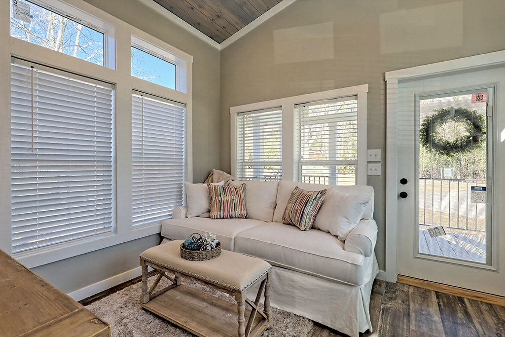 Bright, cozy room with a white sofa adorned with striped cushions. Sunlight streams through blinds on large windows and a glass door with a wreath, creating a warm and inviting atmosphere.