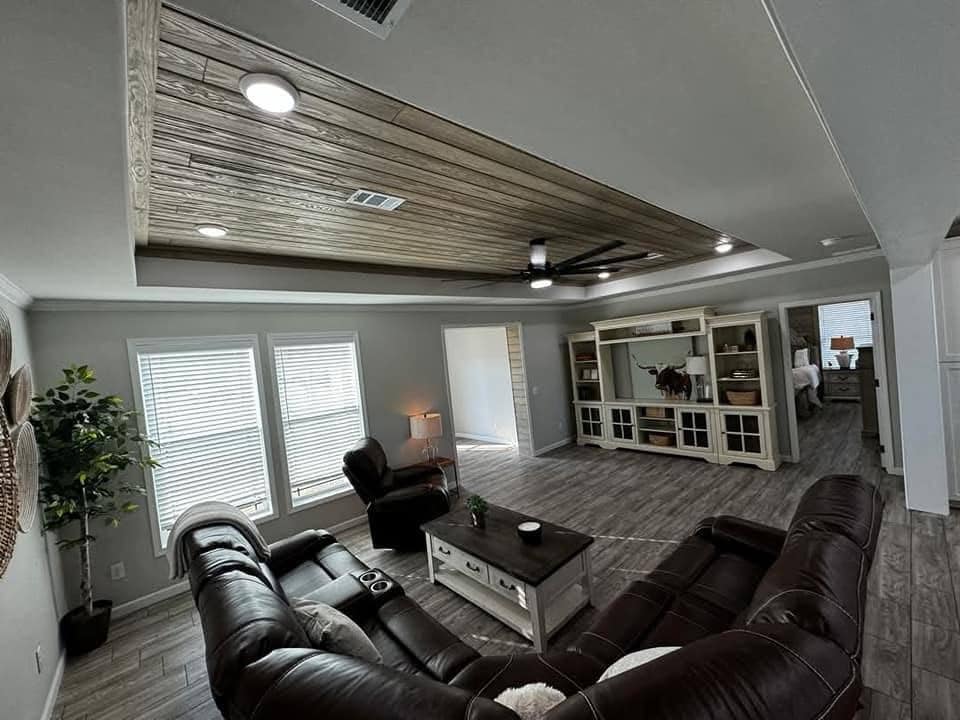 Modern living room with brown leather sectional sofa, rustic wood ceiling, and gray wood flooring. Large windows, entertainment center, and cozy ambiance.