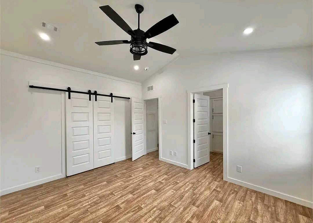 Bright, empty room with light wood flooring, white walls, a black ceiling fan, recessed lighting, and a sliding barn door closet. Modern and spacious.