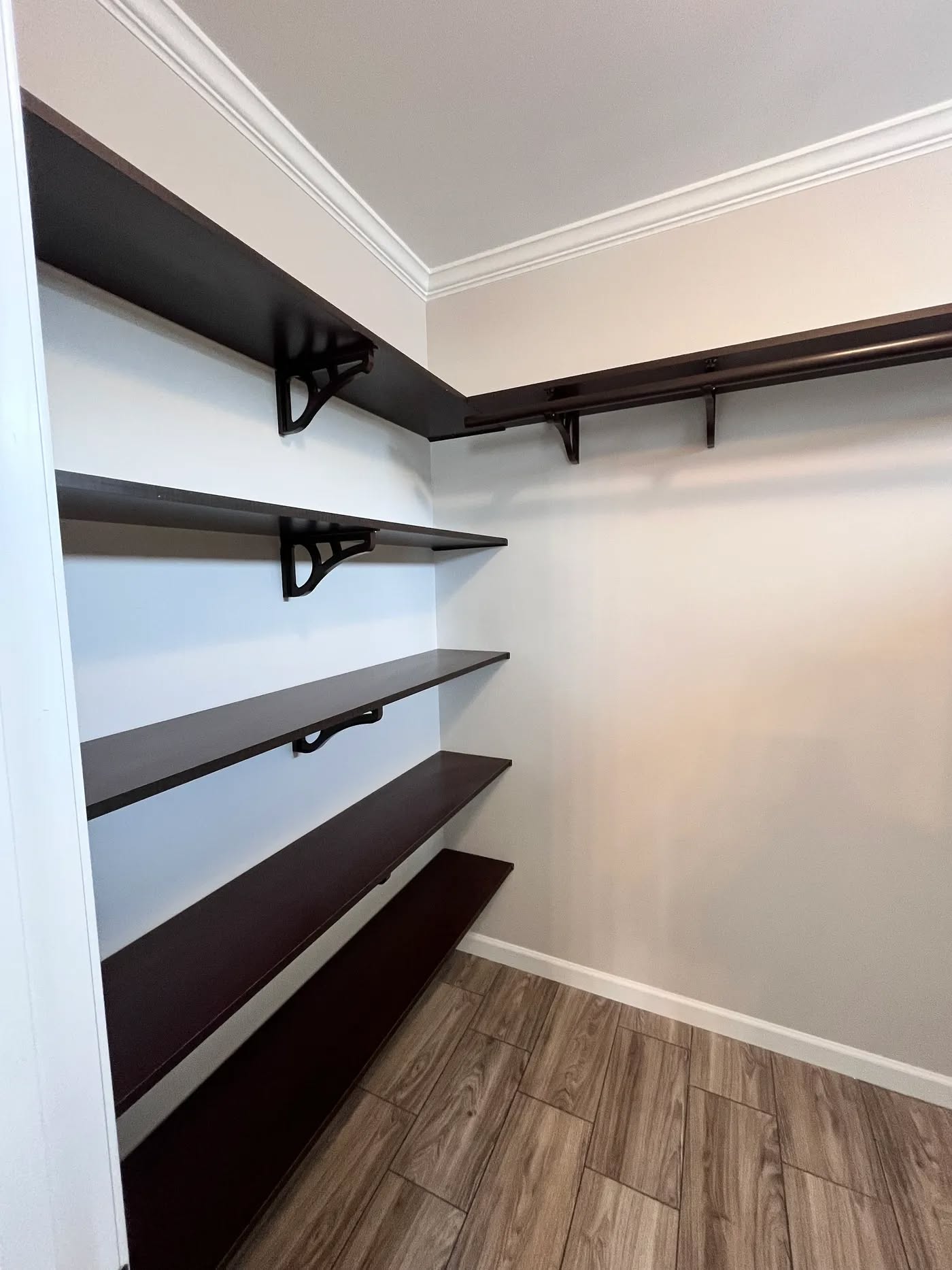 Empty walk-in closet with dark wooden shelves and hanging rod on light-colored walls. Floor features wood-patterned tiles, conveying a modern, clean feel.