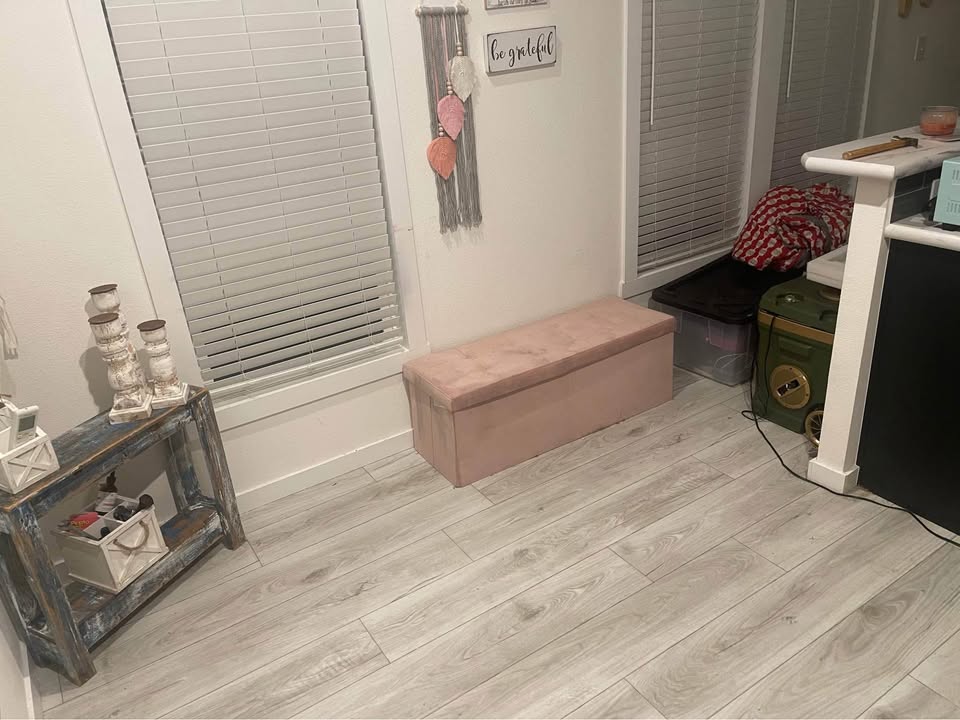 A cozy room corner with a pink cushioned bench, rustic shelf holding candles, and wall decor. Soft lighting gives a warm, inviting feel.