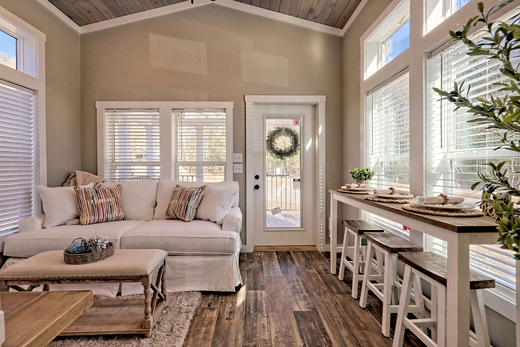 Bright living room with a cozy white sofa, striped pillows, and rustic wood floors. Tall windows and a door with wreath add natural light.