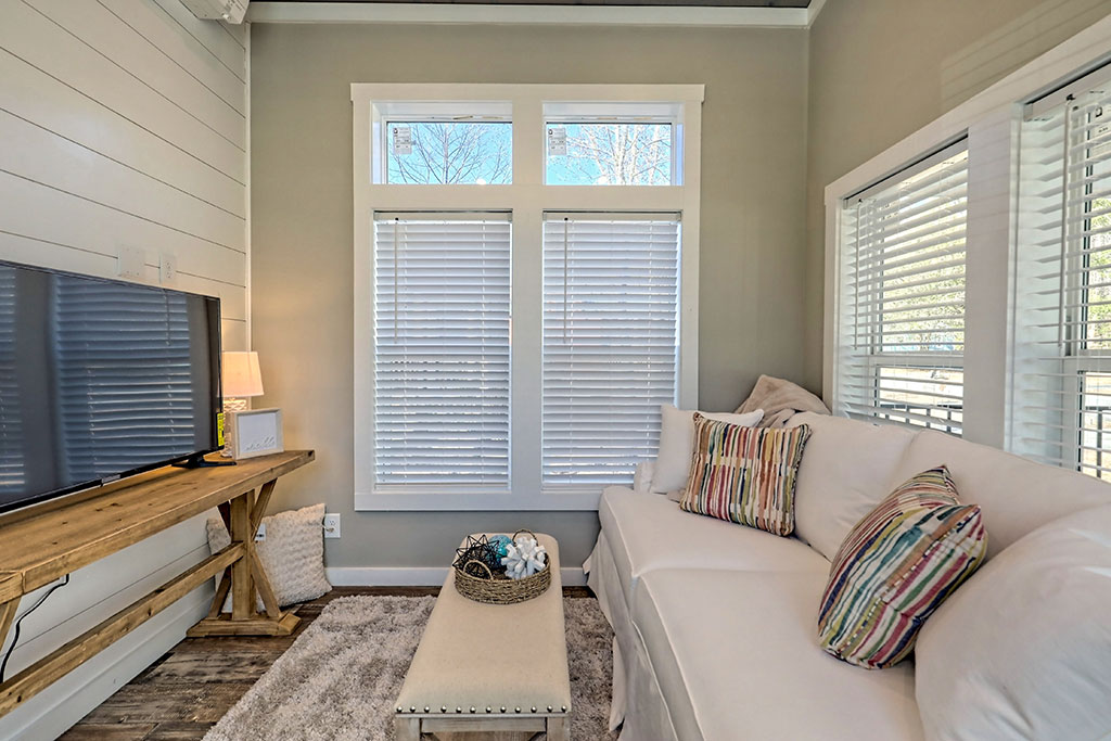 Cozy living room with white sofa adorned with striped cushions, wooden coffee table, and TV on rustic stand. Sunlight filters through blinds, creating a serene ambiance.