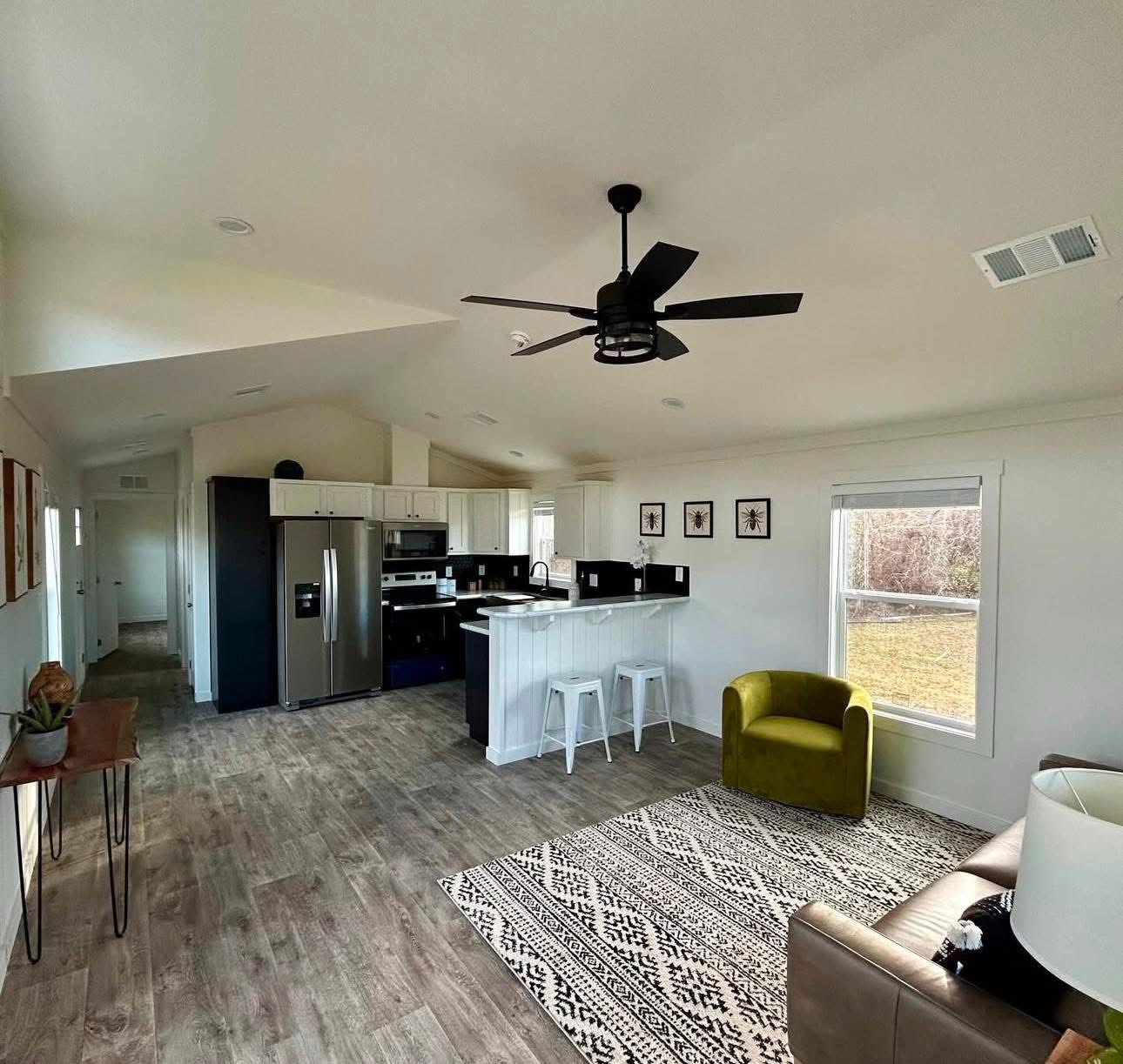 Modern open-plan living area with wood flooring, a cozy olive green chair on a patterned rug, sleek kitchen with stainless steel appliances, and wall art. Warm, inviting atmosphere.