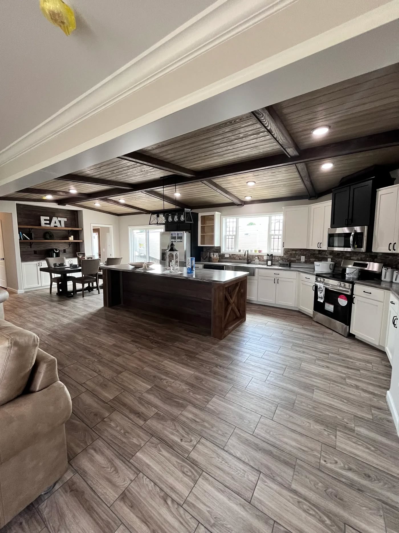 Spacious modern kitchen with dark wood island, white cabinets, stainless steel appliances. Dining area to the left with "EAT" wall decor, warm ambiance.