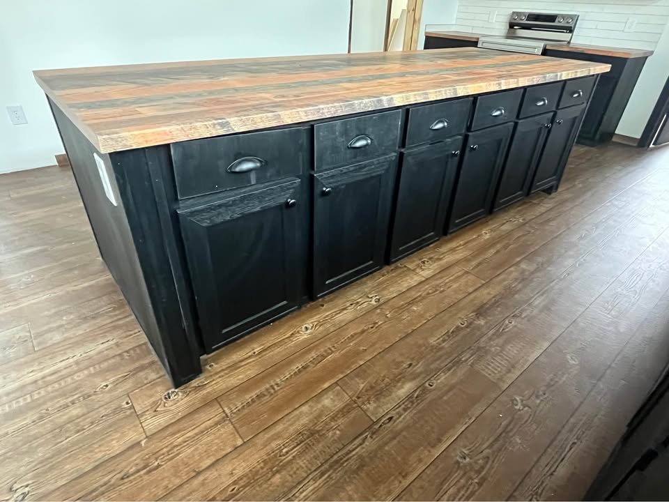 A spacious kitchen island features a rustic wood countertop and black cabinets with sleek handles. It complements the warm-toned wooden flooring.