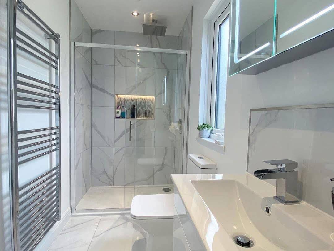 Modern bathroom with marble tiles, glass shower enclosure, and a sleek sink. A large window, chrome towel rack, and small plant add elegance.