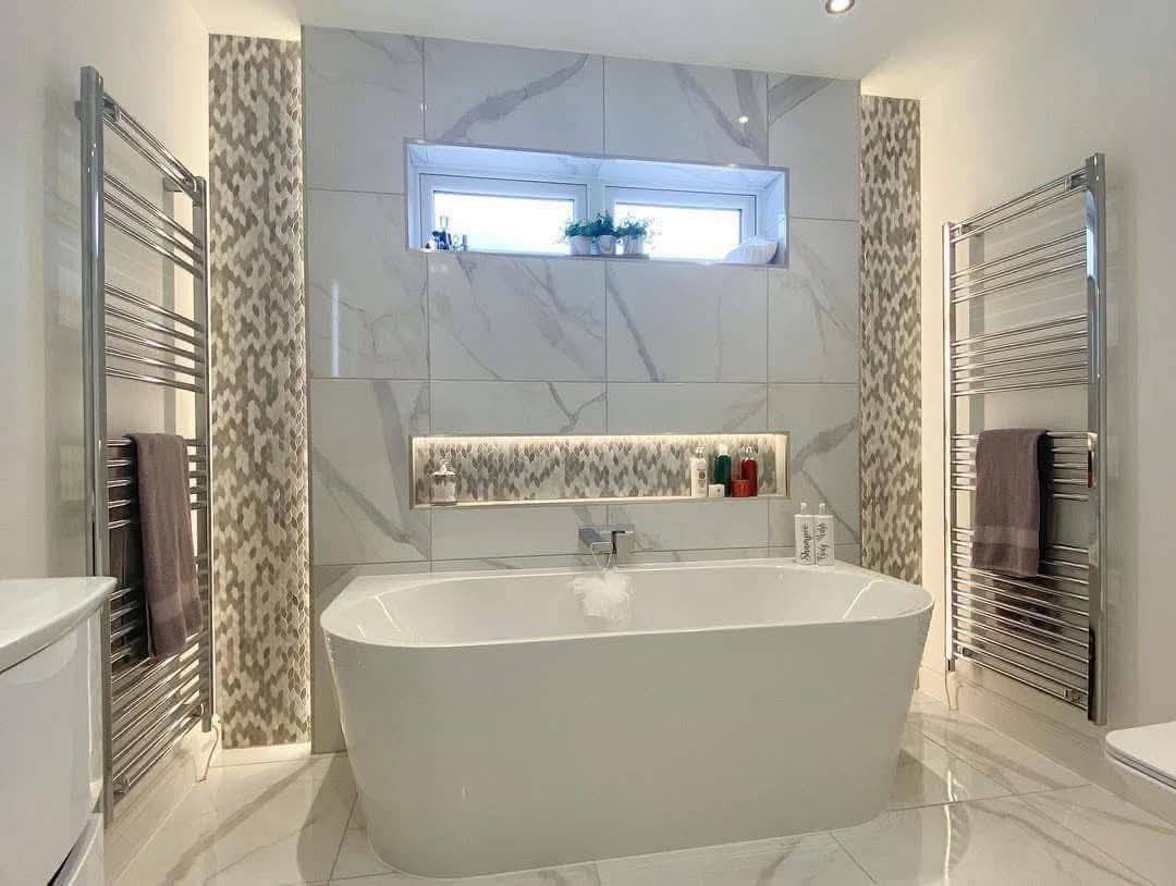 Modern bathroom featuring a freestanding white bathtub, gray marble tiles, dual heated towel racks, small window, and recessed lighting with toiletries.