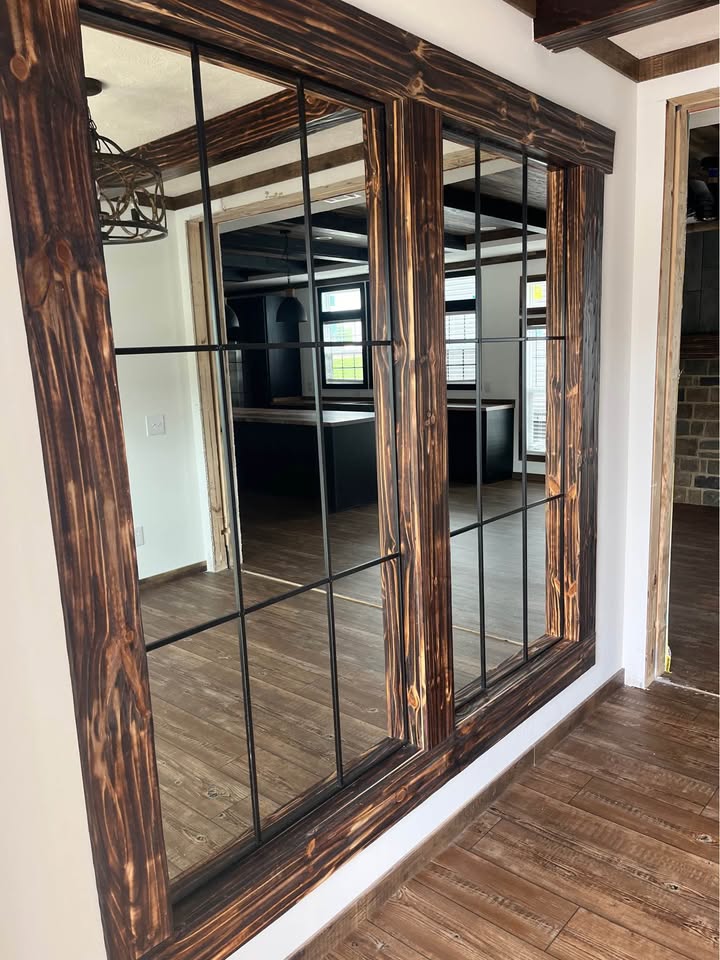 Reflective wall mirrors with rustic wooden frames in a room with wooden floors. The mirrors reflect an adjacent room with windows and brick accents.
