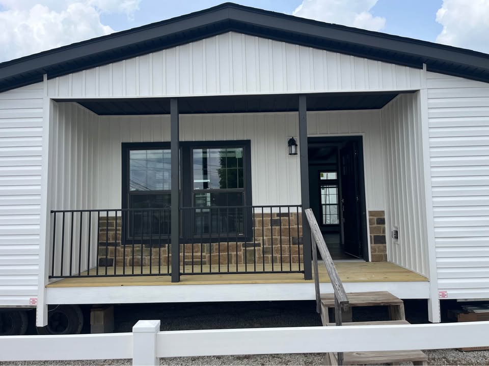 White modular home with a small wooden porch, black railing, and stone accents. A black door and window create a modern, welcoming vibe. Cloudy sky above.