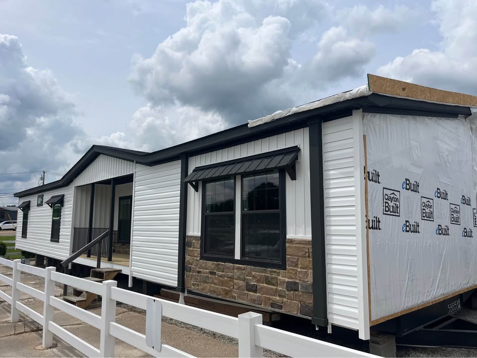 A modern manufactured home with a white and stone exterior, partially wrapped in branded plastic, stands under a cloudy sky. A white fence is in the foreground.