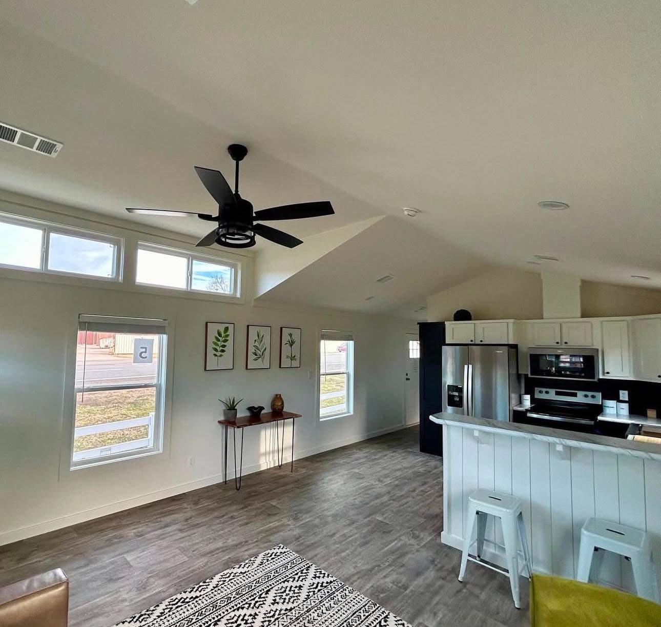 Modern living room and kitchen with high ceilings and a ceiling fan. Features include a patterned rug, bar stools, and framed leaf art. Bright, airy ambiance.
