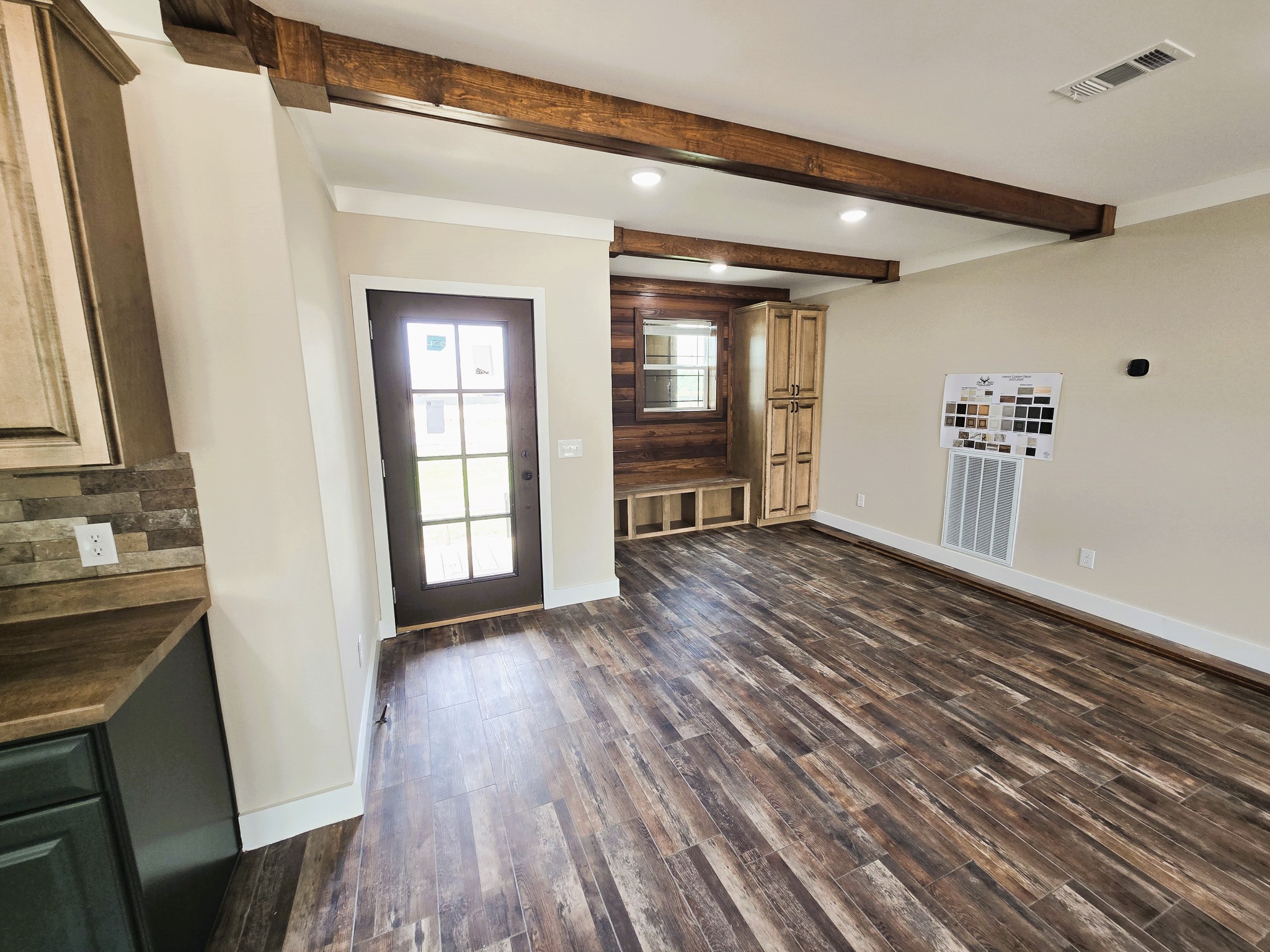 A room with wood flooring, a dark wooden door, exposed ceiling beams, and beige walls. Cabinets and a bench are near the window, creating a cozy feel.