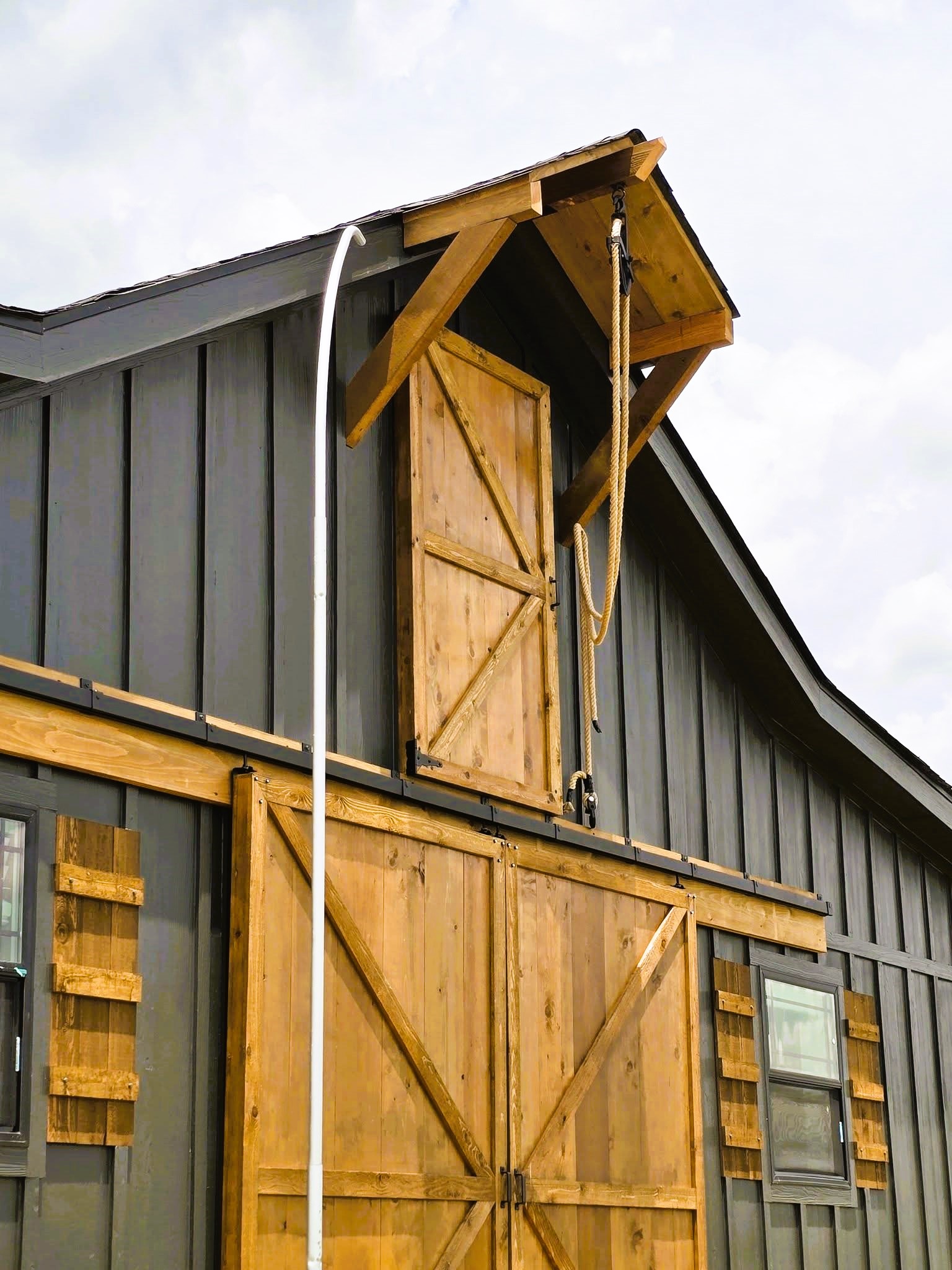 Barn exterior with dark gray siding and wooden details. Features large wooden sliding doors, smaller top door with a pulley, and windows. Rustic and functional.