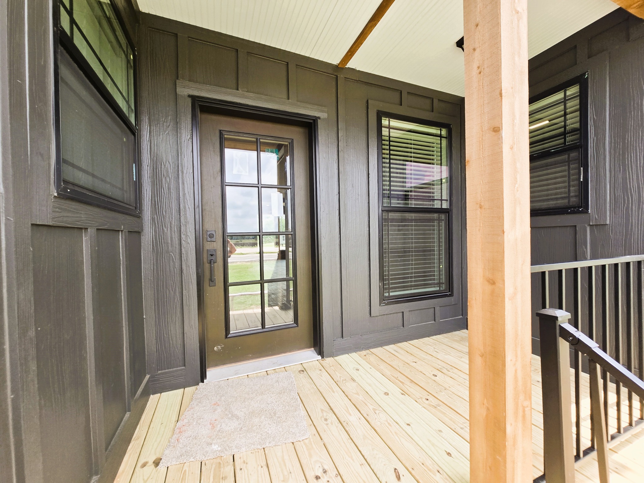 Covered porch with a wooden floor and dark painted walls. Features a glass-paneled door, two large windows with blinds, and a simple doormat.