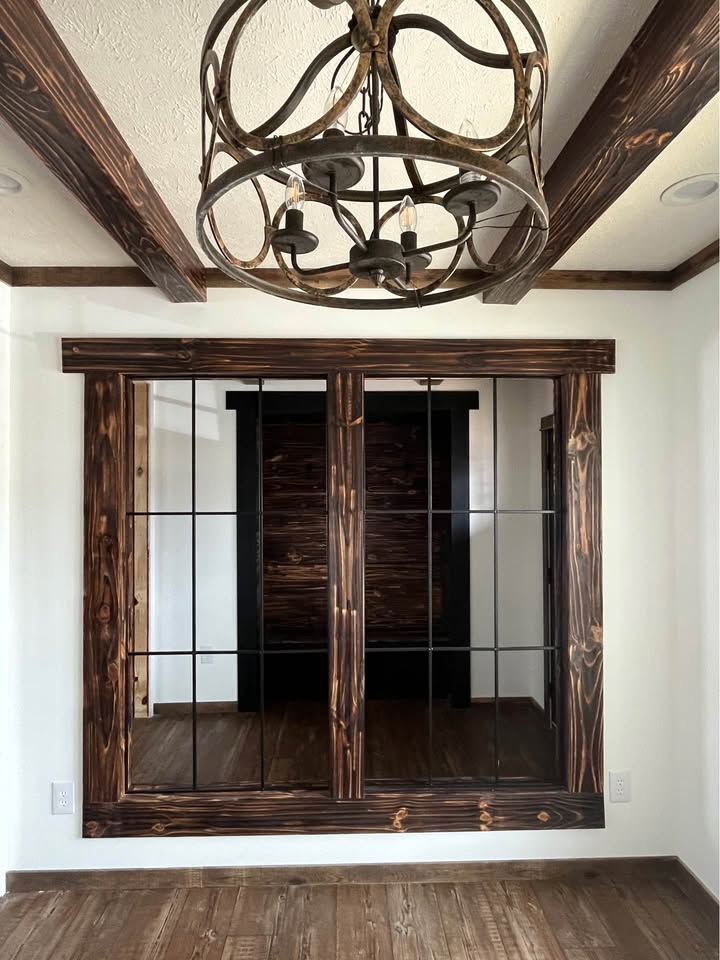 Rustic interior features dark wood beams and a vintage-style black metal chandelier. There's a large window with grid panes and matching wood trim. The atmosphere is cozy and warm.