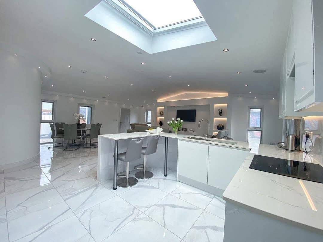 Modern, open-concept kitchen and living room with white marble floors, sleek white cabinets, a skylight, bar stools, and a dining area in the background.