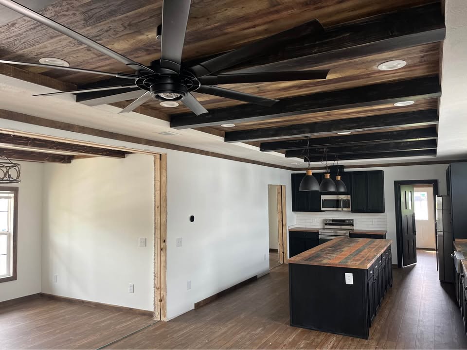 Modern kitchen interior with a rustic feel, featuring dark wood beams, a large ceiling fan, and a central island with wooden countertops. Bright, open space.