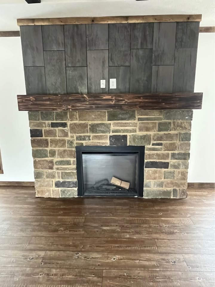 Stone fireplace with a wooden mantel against a dark paneled wall. An empty firebox sits above a hardwood floor, creating a rustic ambiance.