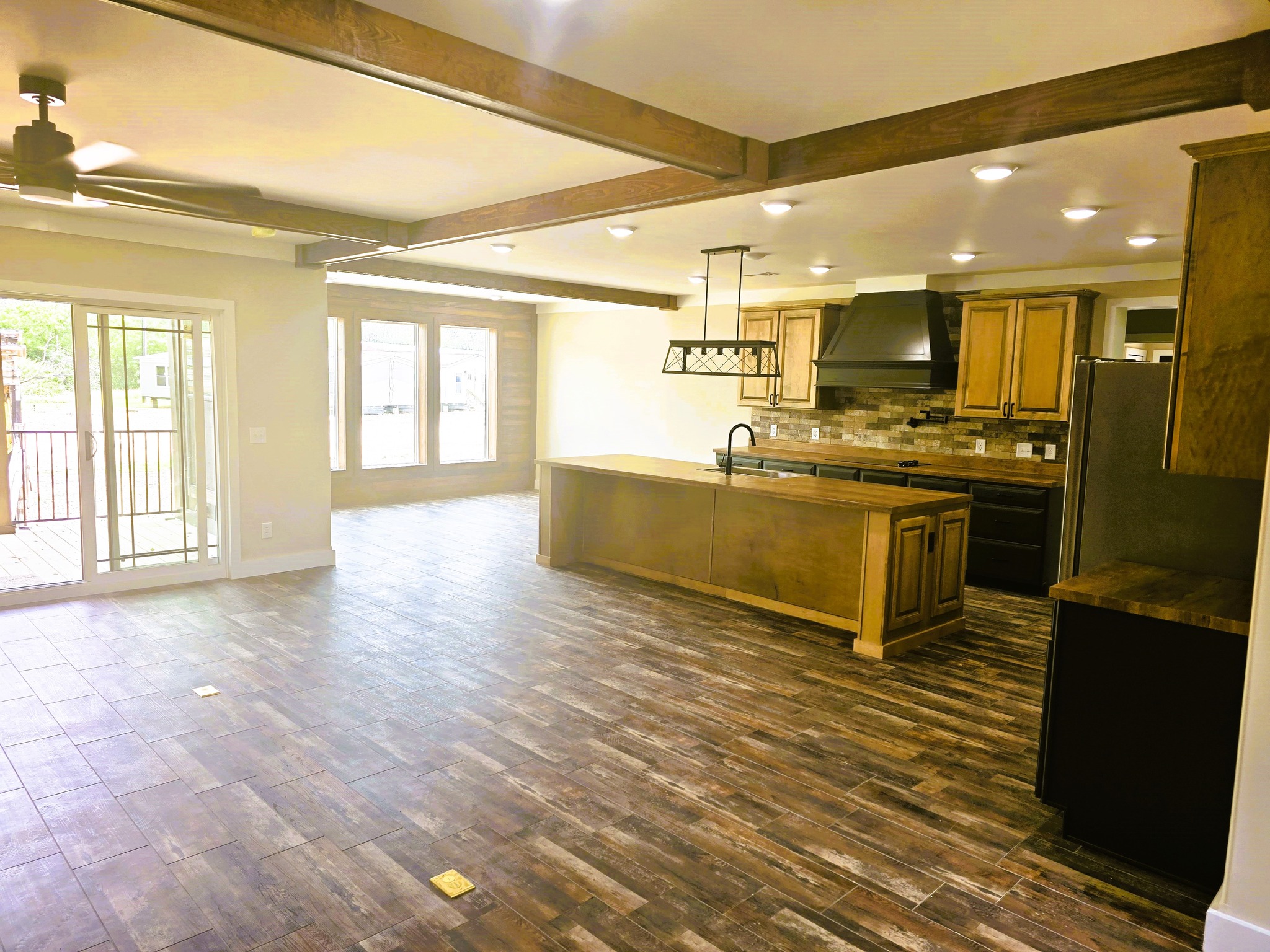 Spacious kitchen with wooden floors and cabinets, black appliances, and large windows. An island with a hanging light exudes a modern, warm ambiance.