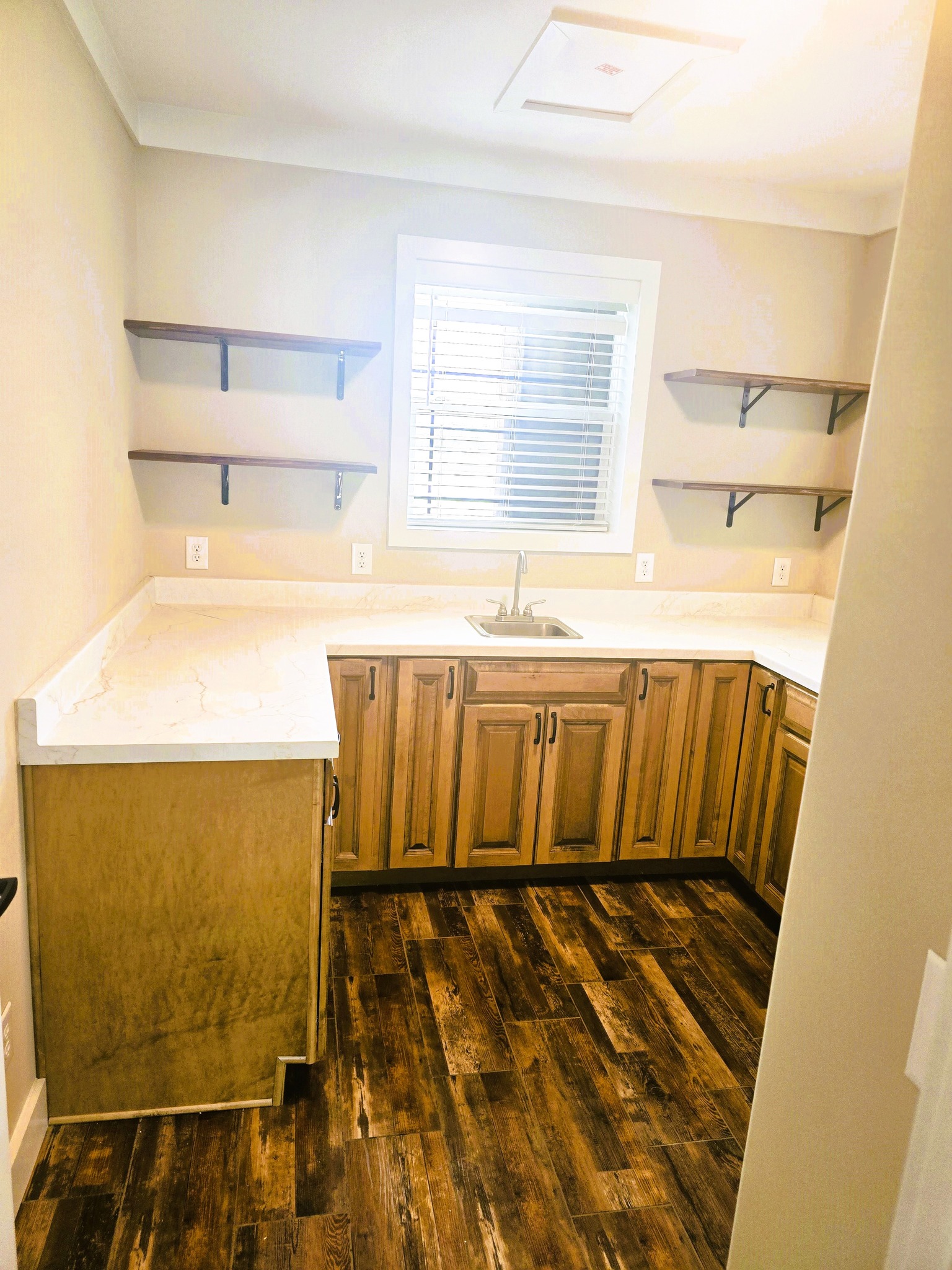 Bright kitchen with light wood cabinets and marble countertops. Dark wood flooring contrasts with open shelves and a window over the sink. Cozy and inviting.