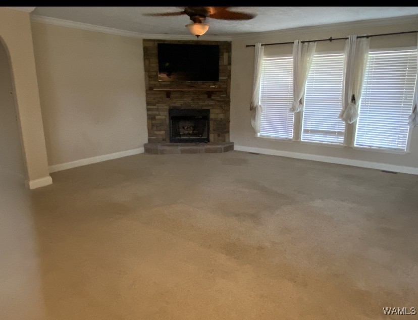 Empty living room with beige carpet, a stone fireplace, and a wall-mounted TV. Large windows with blinds and curtains let in soft natural light. Cozy and inviting atmosphere.