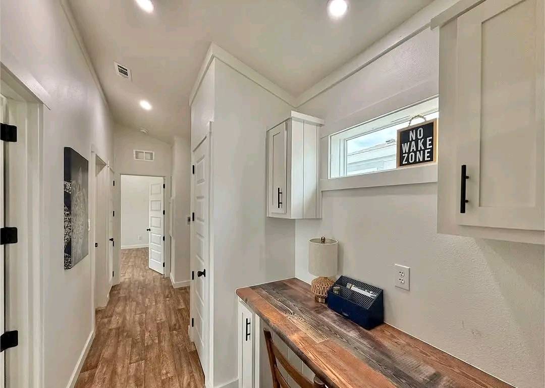 Hallway with wooden floors, leading to a white door. On the right, a small desk with a lamp and a "No Wake Zone" sign above, creating a cozy nook.