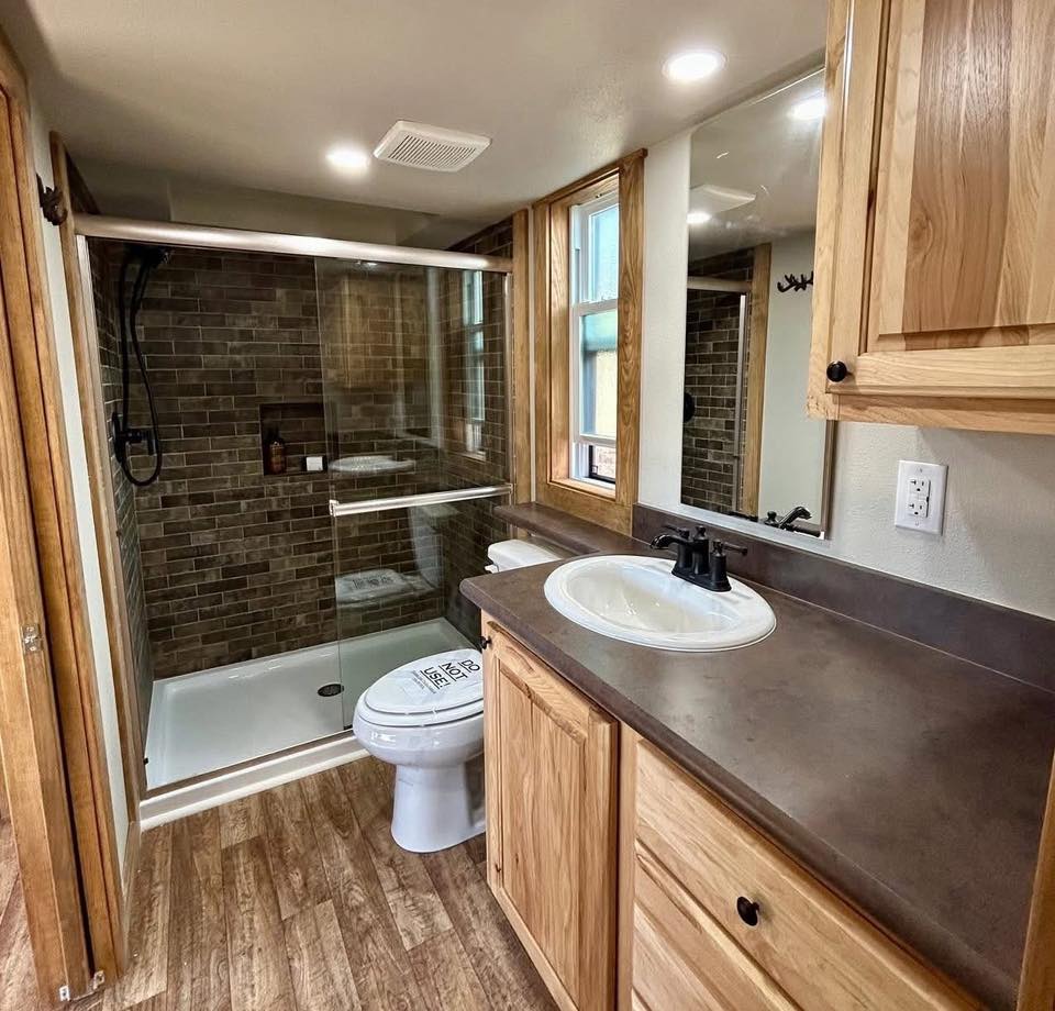 A small bathroom with a wood-finished vanity, white sink, and dark faucet. A glass-enclosed shower with brown tiles is on the left. Warm, natural tones.