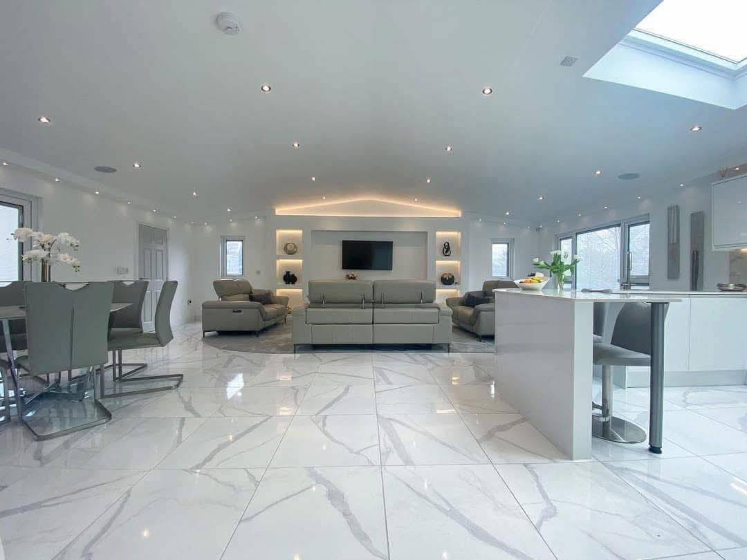 Spacious modern living room with white marble floors, gray sofas, and recessed lighting. A TV is mounted on the back wall. Airy, minimalist feel.