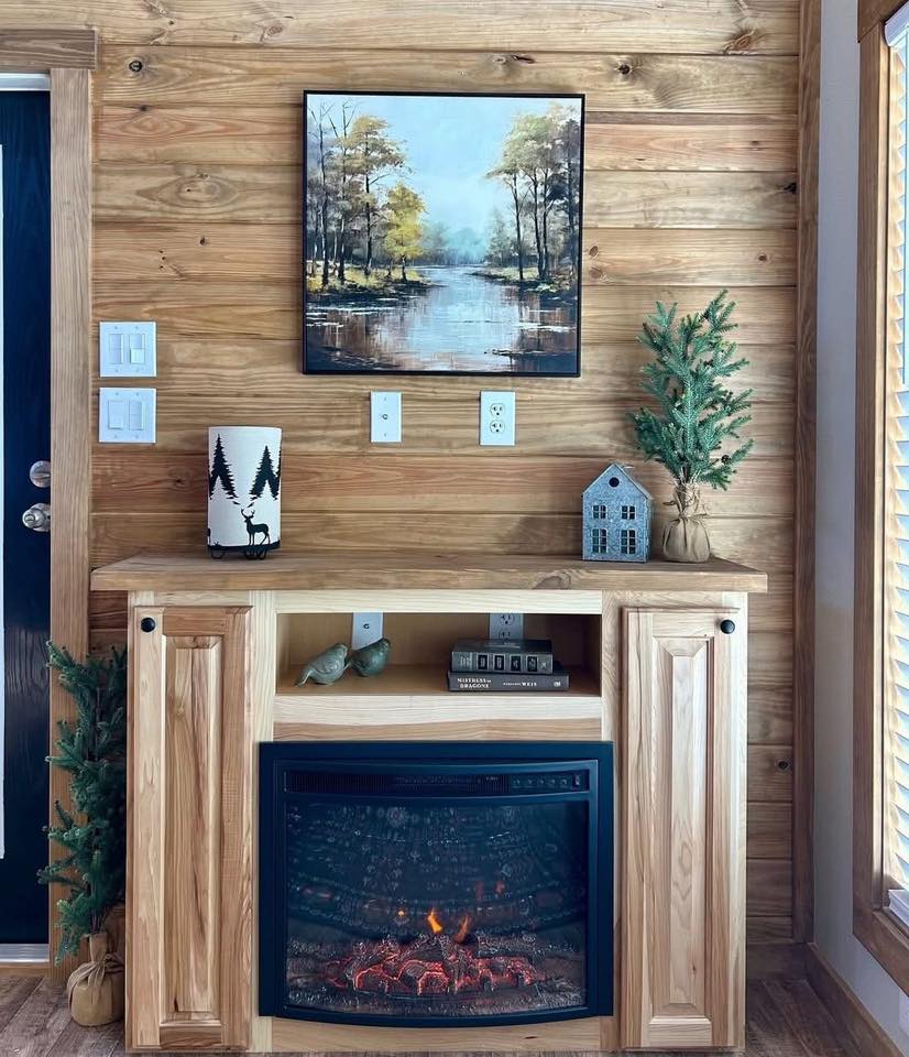 Cozy wooden room with an electric fireplace on a pine mantel. Above, a serene landscape painting features trees and a reflective lake. Rustic decor accents.