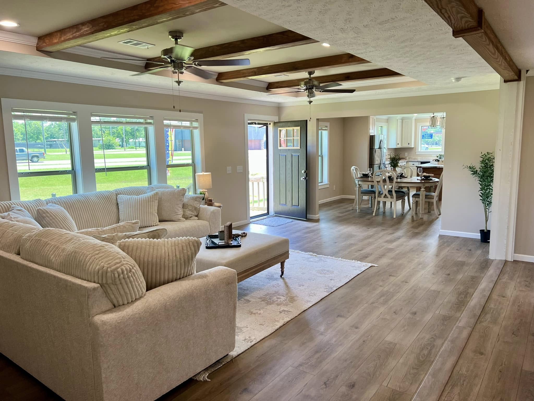 Spacious living room with light wood floors and beige walls. Features a large white sectional sofa, ceiling fans, and beams. Open doorway to dining area.
