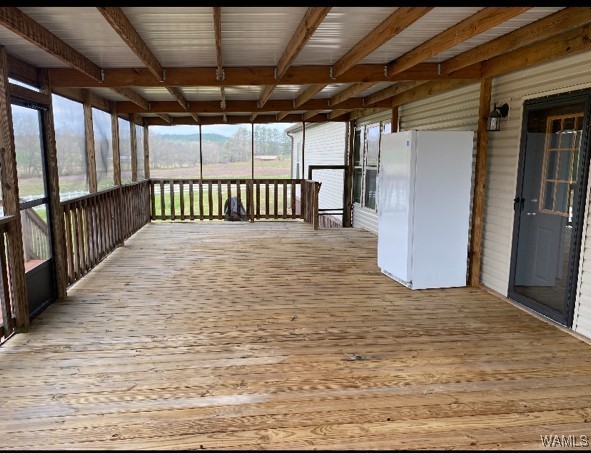 A spacious, screened-in wooden porch with a white refrigerator on the right. Large windows and a door provide natural light; a serene rural landscape is visible.