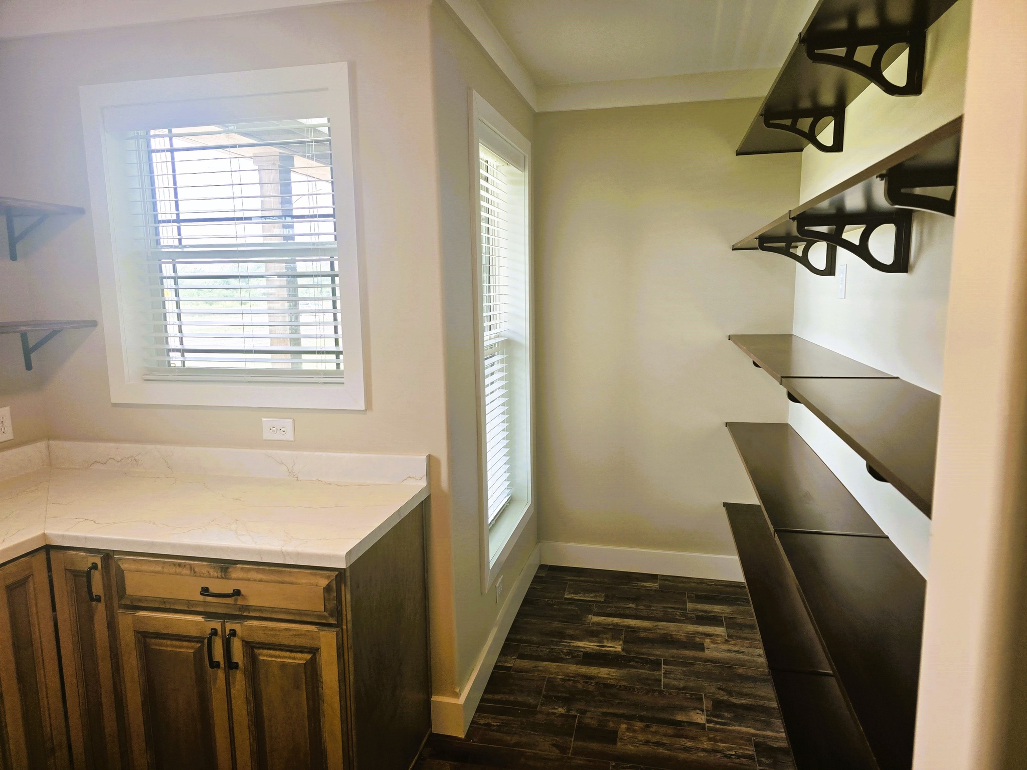 A pantry with dark wooden shelves on the right, light wooden cabinets with a marble countertop on the left, and two sunlit windows enhancing the warm ambiance.