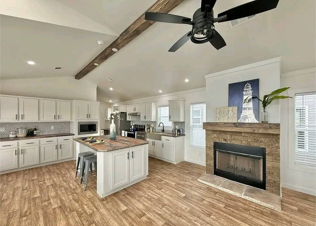 Spacious modern kitchen with white cabinets and wood accents. Features a center island, stone fireplace, ceiling fan, and large windows, creating a bright, cozy atmosphere.