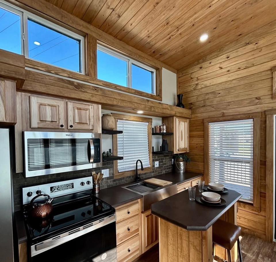 Cozy kitchen with rustic wood paneling and natural light. Features stainless steel appliances, black countertops, two stools, and a small dining area.