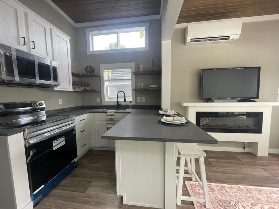 Modern kitchen and living area with white cabinets, a black countertop, and wooden shelves. A TV and electric fireplace add a cozy feel.
