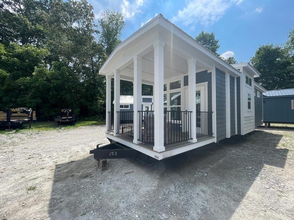 Small grey house with a white porch, black railings, and large windows in a gravel lot, surrounded by trees under a partly cloudy sky. Peaceful vibe.