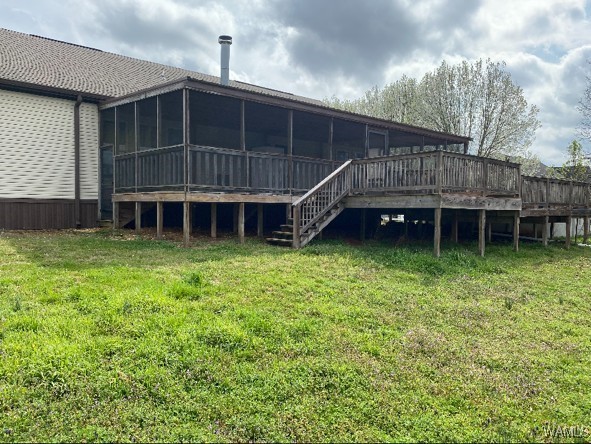 A wooden deck extends from a house with a screened porch and chimney. The deck is elevated with stairs, surrounded by a lush green yard under a cloudy sky.