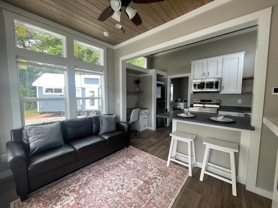 Cozy living area in a tiny house with large windows, brown sofa, and a patterned rug. Adjacent kitchen features white cabinets and bar stools, exuding a calm, modern vibe.