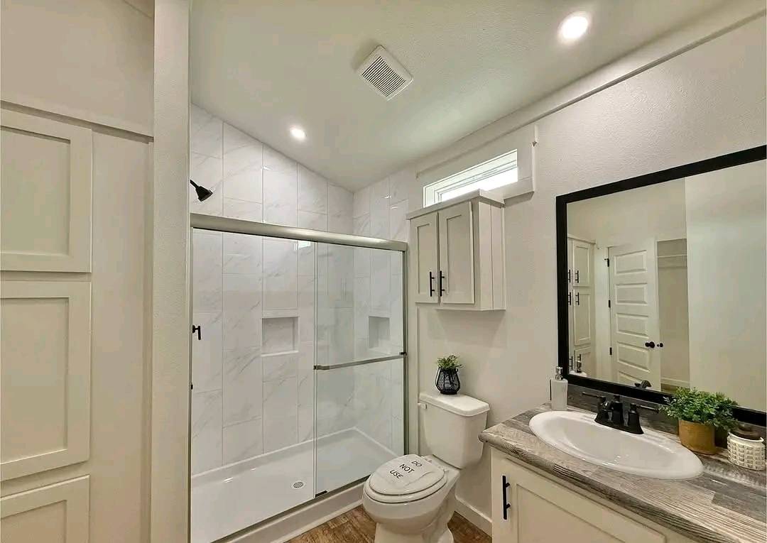Modern bathroom with a sleek white design. Features a glass shower, toilet, large mirror, and sink on a wooden countertop. Bright, clean, and inviting.