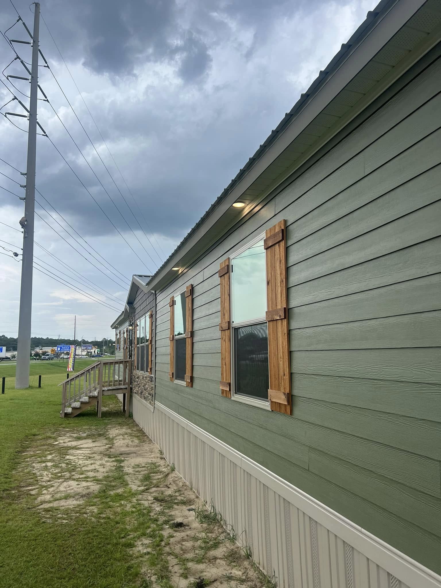A green house with wooden shutters under a cloudy sky, with a grassy yard and power lines nearby, conveying a calm and rustic feel.